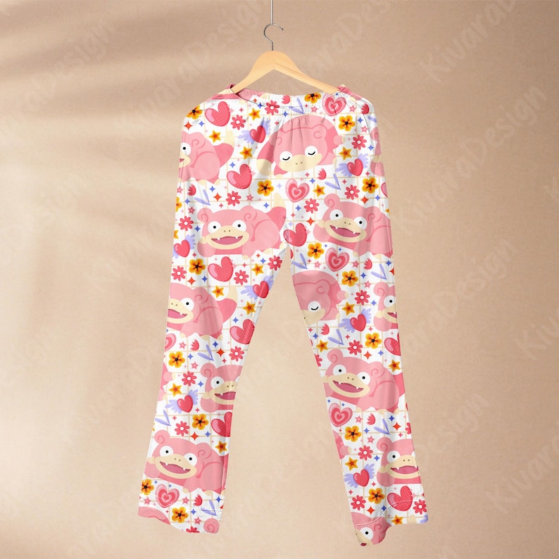 K&ouml;nnte beinhalten: Rosa Schlafanzughose mit einem sich wiederholenden Muster aus Zeichentrickfiguren, Herzen und Blumen. Die Hose h&auml;ngt an einem Holzkleiderb&uuml;gel vor einem neutralen Hintergrund. Die Schlafanzughose ist aus einem weichen, leichten Stoff gefertigt.