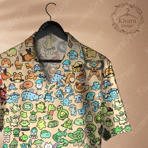 May include: A short-sleeved button-down shirt with a beige background and a colorful cartoon character pattern. The shirt is hanging on a black hanger. The shirt features a variety of creatures in shades of blue, green, orange, and yellow.