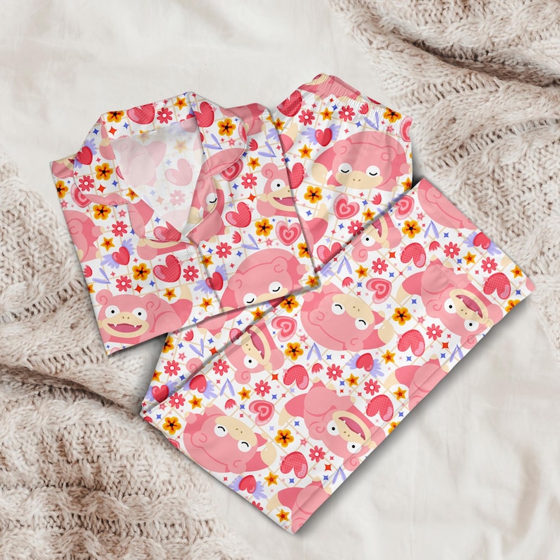 K&ouml;nnte beinhalten: Rosa Pyjama-Set mit einem sich wiederholenden Muster aus Zeichentrickfiguren, Herzen und Blumen auf wei&szlig;em Hintergrund. Das Pyjama-Oberteil hat einen Kragen und einen Knopfverschluss. Das Set enth&auml;lt ein lang&auml;rmliges Oberteil und eine passende Hose.
