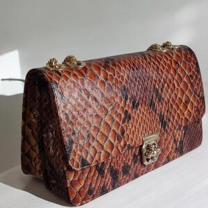 May include: A brown snakeskin-patterned handbag with a gold chain strap and a gold clasp.
