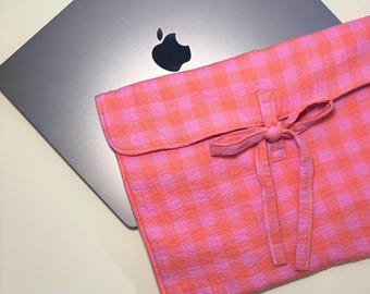 May include: A silver laptop with a black Apple logo is partially visible. A pink and orange gingham laptop sleeve with a matching tie closure is in the foreground. The sleeve is made of a textured fabric.