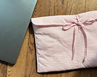 May include: A pink and white gingham laptop sleeve with a matching ribbon tie closure. The sleeve is made of fabric and is designed to protect a laptop. A silver laptop is partially visible.