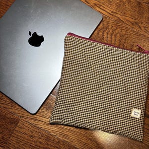 May include: A silver laptop with a black Apple logo rests on a wooden surface next to a small, square pouch. The pouch is patterned with a brown and tan gingham check and has a burgundy zipper. A small white tag reads "birdie bear."
