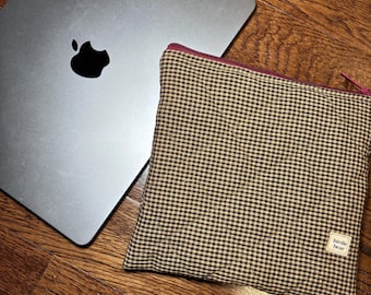 May include: A silver laptop with a black Apple logo rests on a wooden surface next to a small, square pouch. The pouch is patterned with a brown and tan gingham check and has a burgundy zipper. A small white tag reads "birdie bear."