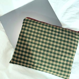 May include: A green and tan plaid pattern laptop sleeve with a red zipper. The sleeve is resting on top of a silver laptop, with a white background. The sleeve is made of fabric and is rectangular in shape.