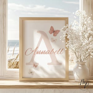 May include: A framed print featuring a large pink letter "A" with the name "Annabell" in script. Pink butterflies are scattered around the letter. The print is in a light wood frame, with a beach scene visible in the background.