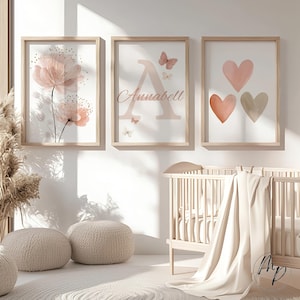 May include: Three framed art prints with a nursery theme. One print features pink flowers, another has the letter "A" with the name "Annabell" and butterflies, and the third displays three hearts in shades of pink and beige. The frames are light wood.