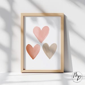 May include: A framed art print featuring three watercolor hearts in shades of pink, peach, and taupe. The hearts are arranged in a vertical composition against a white background. The frame is made of light-colored wood.