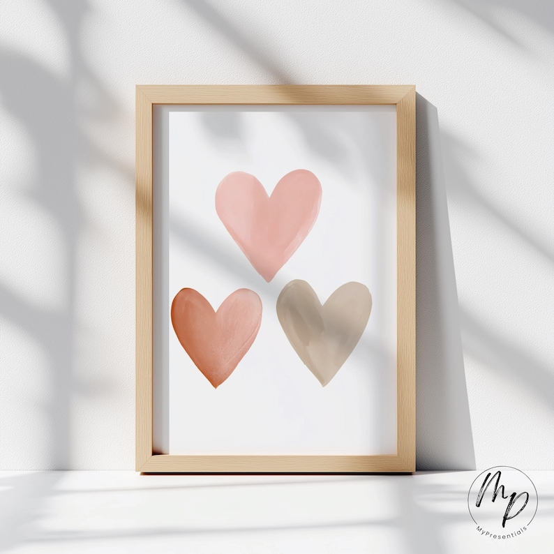 May include: A framed art print featuring three watercolor hearts in shades of pink, peach, and taupe. The hearts are arranged in a vertical composition against a white background. The frame is made of light-colored wood.