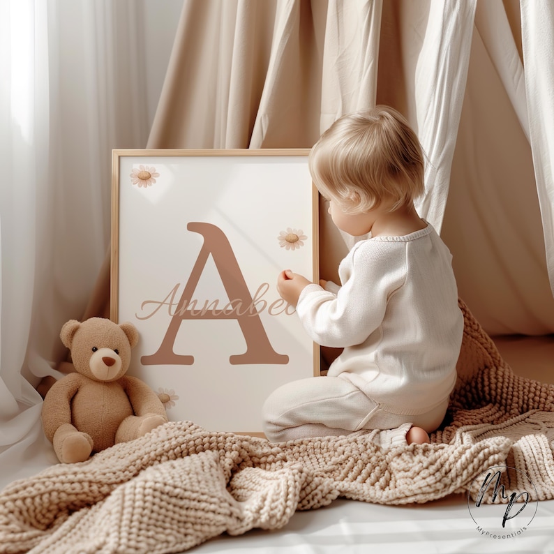 May include: A framed print featuring a large letter "A" and the name "Annabel" in a soft brown hue, adorned with daisy accents. A teddy bear and a child's blanket are in the foreground. The child is wearing a white outfit.