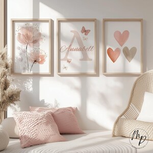 May include: Three framed art prints with a soft, feminine aesthetic. One features pink flowers, another a letter "A" with the name "Annabell" and butterflies, and the third, three hearts in shades of pink and beige. The frames are light wood.