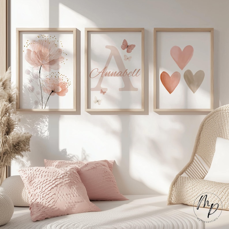 May include: Three framed art prints with a soft, feminine aesthetic. One features pink flowers, another a letter "A" with the name "Annabell" and butterflies, and the third, three hearts in shades of pink and beige. The frames are light wood.