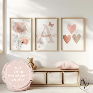 Könnte beinhalten: Drei gerahmte Kunstdrucke mit einem Blumendesign, dem Buchstaben "A" mit dem Namen "Annabell" und drei Herzen in Rosa- und Beigetönen. Die Drucke werden über einer Holzbank mit Aufbewahrungswürfeln angezeigt. Ein Teddybär und ein rosa Kissen sind ebenfalls sichtbar.