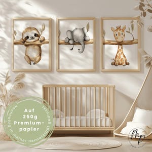Könnte beinhalten: Drei gerahmte Aquarelldrucke mit einem Faultier, einem Elefanten und einer Giraffe auf Ästen. Die Drucke sind in hellen Holzrahmen. Eine Holzkinderbett und ein Hängesessel mit einem weißen Kissen befinden sich im Vordergrund. Der Text auf dem Bild lautet "Auf 250g Premium-papier".