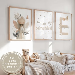 Könnte beinhalten: Drei gerahmte Kunstdrucke mit hellen Holzrahmen. Einer zeigt eine Fee, ein anderer sagt "You are my sunshine", und der dritte hat den Namen "Emma". Die Drucke befinden sich über einem Bett mit Teddybären und Kissen.