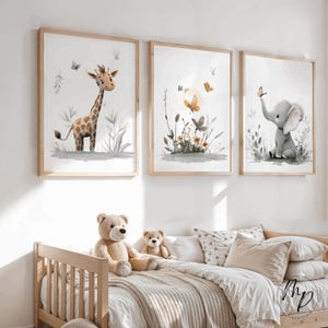 K&ouml;nnte beinhalten: Drei gerahmte Aquarelldrucke mit einer Giraffe, Schmetterlingen und einem Elefanten. Die Drucke haben einen hellen Holzrahmen und werden &uuml;ber einem Kinderbett mit Teddyb&auml;ren und Kissen ausgestellt. Das Kunstwerk hat eine weiche, neutrale Farbpalette.