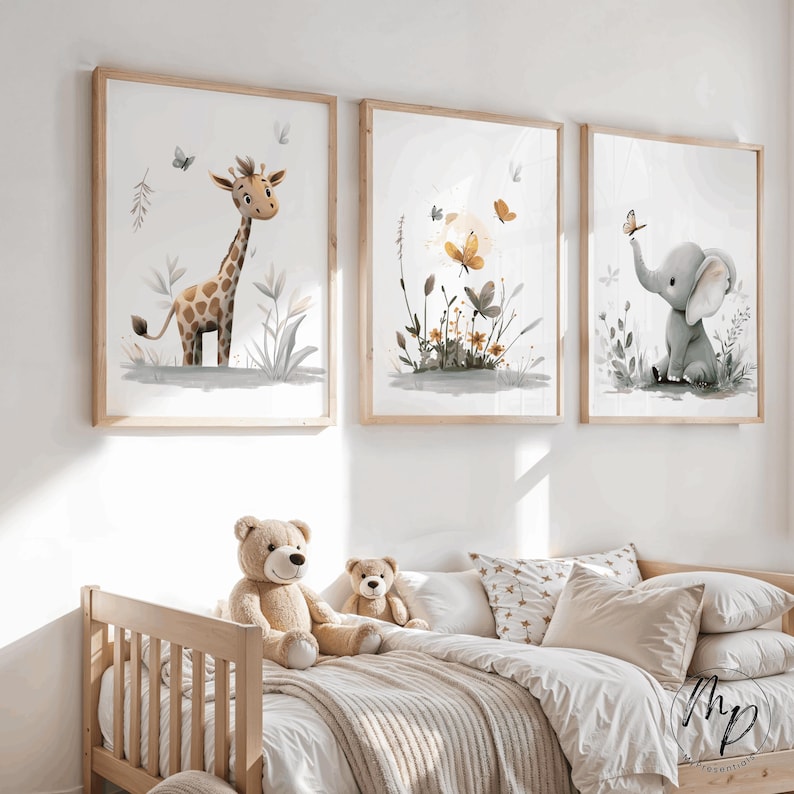 K&ouml;nnte beinhalten: Drei gerahmte Aquarelldrucke mit einer Giraffe, Schmetterlingen und einem Elefanten. Die Drucke haben einen hellen Holzrahmen und werden &uuml;ber einem Kinderbett mit Teddyb&auml;ren und Kissen ausgestellt. Das Kunstwerk hat eine weiche, neutrale Farbpalette.