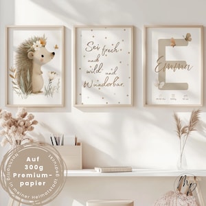 Può includere: Tre stampe artistiche incorniciate. Una presenta un riccio con una corona floreale, un'altra ha il testo "Sei frech und wild und Wunderbar", e la terza mostra il nome "Emma" con dettagli di nascita. Le stampe sono in tonalità neutre.