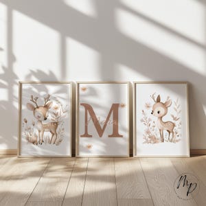 May include: Three framed art prints featuring a woodland theme. Each print has a light wood frame. Two prints depict a fawn surrounded by flowers. The center print displays the letter "M" and the name "Maria".