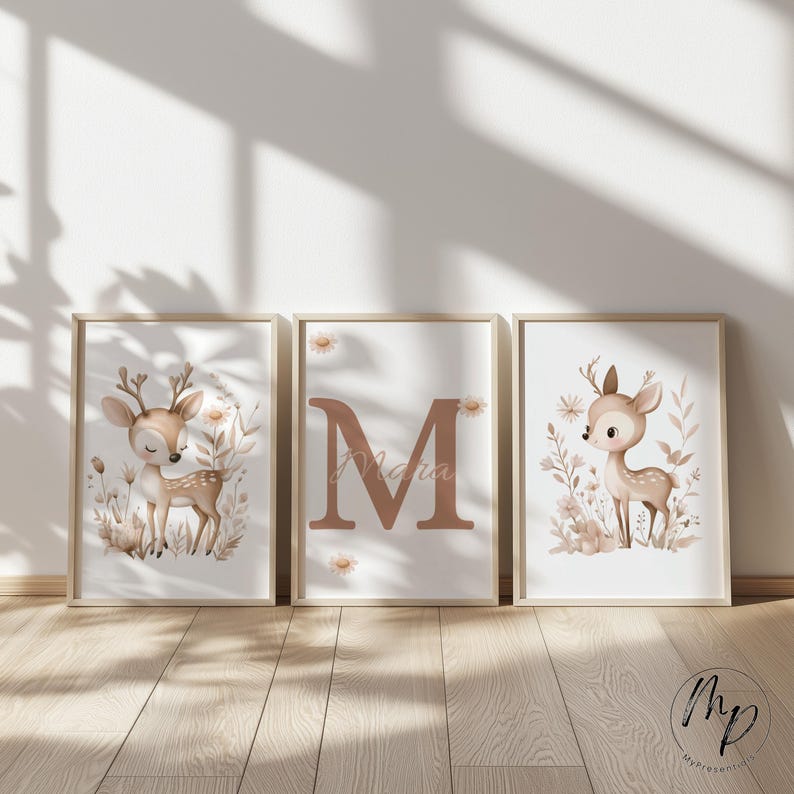 May include: Three framed art prints featuring a woodland theme. Each print has a light wood frame. Two prints depict a fawn surrounded by flowers. The center print displays the letter "M" and the name "Maria".