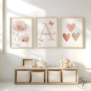 May include: Three framed art prints with a floral design, the letter "A" with the name "Annabell", and three hearts. The prints are in soft pink and beige tones, with wooden frames. A wooden storage unit with fabric bins and decorative items is below.