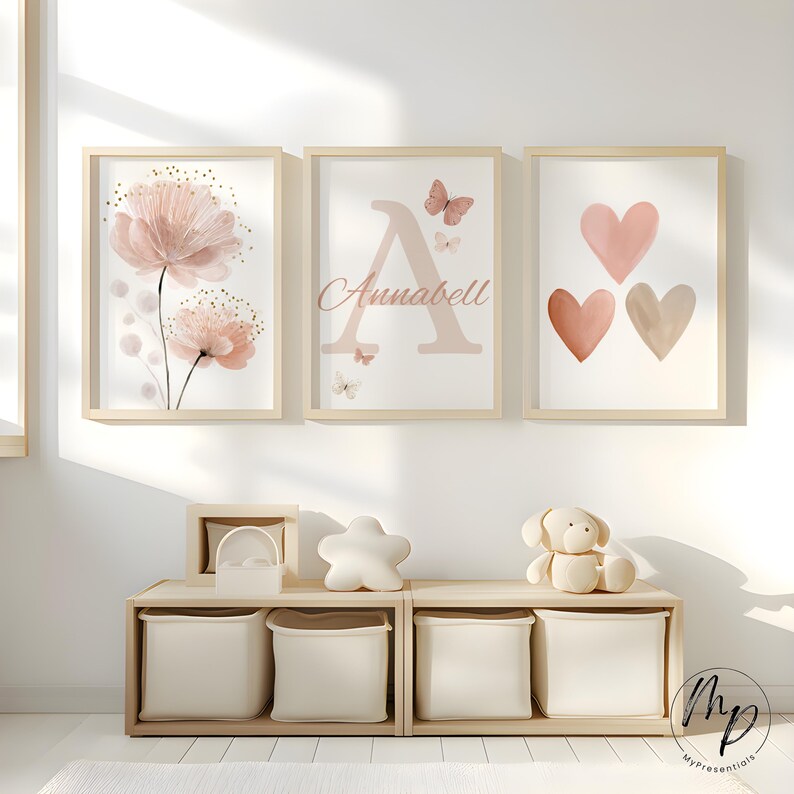 May include: Three framed art prints with a floral design, the letter "A" with the name "Annabell", and three hearts. The prints are in soft pink and beige tones, with wooden frames. A wooden storage unit with fabric bins and decorative items is below.