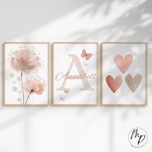 May include: Three framed prints with a floral, initial, and heart theme. The left print features pink watercolor flowers. The center print has the initial "A" and the name "Annabell". The right print displays three hearts in varying shades of pink and beige.