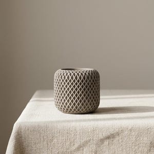 May include: A cylindrical light gray vessel featuring a repeating diamond-shaped lattice cutout pattern across its exterior surface. The object has a smooth, rounded top edge and a solid base, presenting a consistent matte finish throughout its structure.