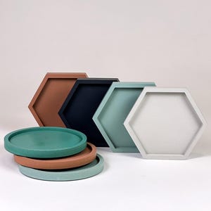 May include: A collection of decorative trays in various colors and shapes. The image features several hexagonal trays in shades of brown, navy, teal, and gray, alongside a stack of round trays in matching colors. The trays are arranged on a white surface.