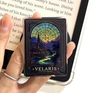 May include: A rectangular phone grip with a stained-glass window design depicting a castle, river, and mountains. The grip has the text "VELARIS - CITY OF STARLIGHT" in gold. The background is a book.