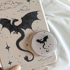 以下が含まれることがあります： ベージュ色のタブレットケースと、黒いドラゴンのデザインの丸い電話グリップ。 ドラゴンは翼を広げ、星と雲に囲まれています。 電話グリップは、ケースのドラゴンのデザインを反映しています。