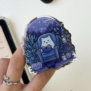 Pode incluir: Um autocolante extravagante com um gato branco a ler um livro numa poltrona azul. A cena passa-se sob um céu estrelado, rodeada de plantas, abóboras e uma pequena mesa. O autocolante tem um acabamento brilhante.