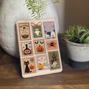 May include: A rectangular Halloween-themed decorative item featuring postage stamp-style illustrations. The stamps depict a potion bottle, ghost, pumpkin, spider, black cat, and haunted house. The item is beige with a decorative bow.