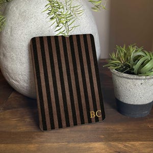 May include: A rectangular notebook with brown and black vertical stripes and gold-coloured initials "BC" in the lower right corner. The notebook is on a wooden surface, with a large white vase and small potted plants in the background. The text "xArtDesignGifts" is visible.