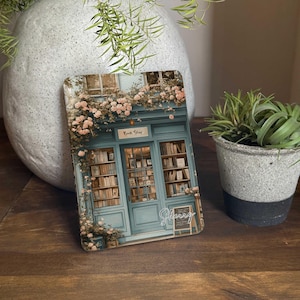 Puede incluir: Una fachada de librería de estilo vintage con una puerta y ventanas de color verde azulado. La fachada está adornada con flores rosas y blancas y el letrero sobre la puerta dice "Book Store".