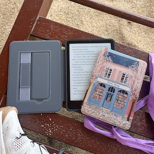 May include: A Kindle e-reader in a grey case, open on a wooden bench. The screen displays text. A decorative cover with a bookstore illustration is visible. The cover has the text "Book Shop" and "Sherry's Kindle".