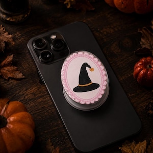 May include: A black smartphone with a circular phone grip attached to the back. The grip features a white oval center with a scalloped pink border, displaying a black witch hat illustration with a gold band and a small gold star at the tip.