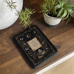 May include: A black e-reader case featuring a gold and white illustration of a crescent moon, butterflies, flowers, and a stack of books. The center displays a parchment-style graphic with the text: Some stories whisper. Some sparkle. Some change everything.