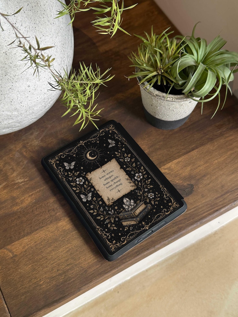 May include: A black e-reader case featuring a gold and white illustration of a crescent moon, butterflies, flowers, and a stack of books. The center displays a parchment-style graphic with the text: Some stories whisper. Some sparkle. Some change everything.