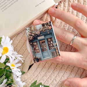 May include: A rectangular phone grip featuring a miniature bookstore scene with a blue facade, floral accents, and the text "Book Shop." The grip is held by a hand, with a book and flowers in the background.