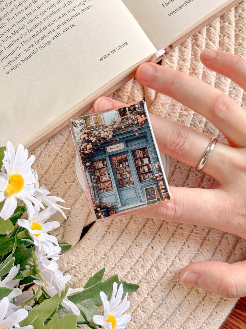 May include: A rectangular phone grip featuring a miniature bookstore scene with a blue facade, floral accents, and the text "Book Shop." The grip is held by a hand, with a book and flowers in the background.