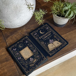 Può includere: Una custodia per e-reader blu scuro con una mezzaluna dorata, farfalle e motivi floreali. Il lato destro mostra un libro aperto con il testo ENCHANTED LIBRARY Where Magic Begins. Il lato sinistro mostra una pergamena con il testo Some stories whisper. Some sparkle. Some change everything.