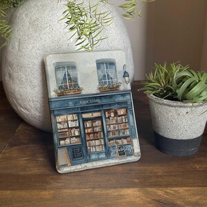 Può includere: Un'illustrazione blu e bianca di una libreria con le parole "Book Store" sopra l'ingresso. La vetrina ha un cartello che dice "Sherry" e una vetrina di libri.