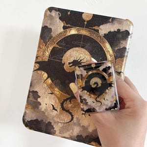 May include: A black and gold decorative case and phone grip featuring a dragon and celestial design. The case has a detailed illustration of a dragon, clouds, and a circular design. The phone grip mirrors the case design.