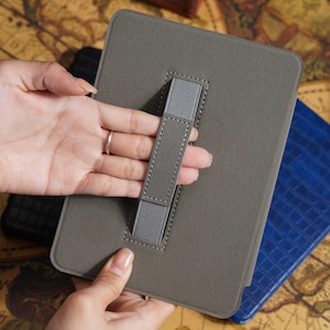 May include: A gray tablet case with a hand strap. The case is rectangular with rounded corners and a stitched border. A gray elastic strap is attached to the back for holding. A blue case is visible in the background.