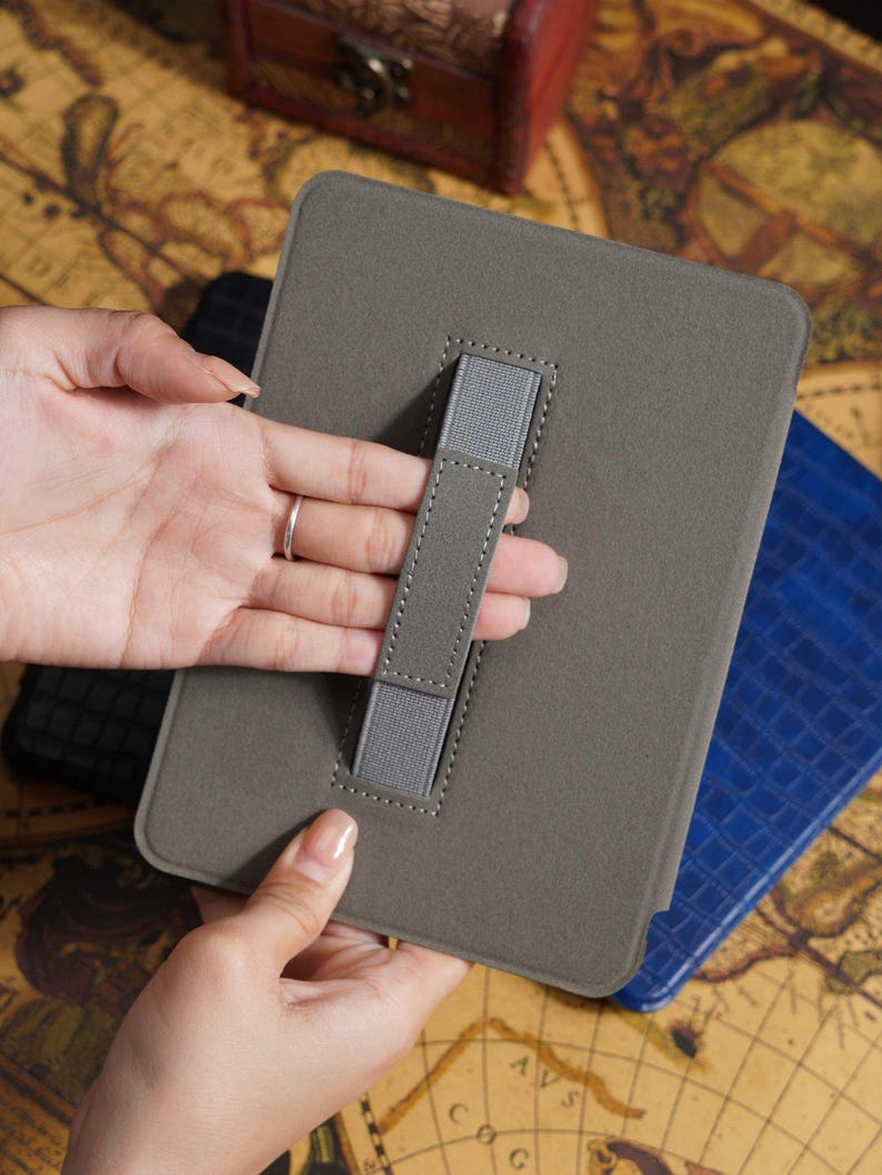 May include: A gray tablet case with a hand strap. The case is rectangular with rounded corners and a stitched border. A gray elastic strap is attached to the back for holding. A blue case is visible in the background.