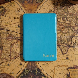 May include: A turquoise leather-bound journal with the name "Karen" in gold lettering. The journal is set on a vintage world map, with a small wooden chest in the background. The journal is rectangular and appears to be closed.