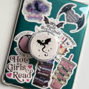May include: A white PopSocket with a black dragon design, surrounded by stars, is attached to a teal phone case covered in stickers. The stickers feature bats, books, and text like "Hot Girls Read" and "My Book Boyfriends."