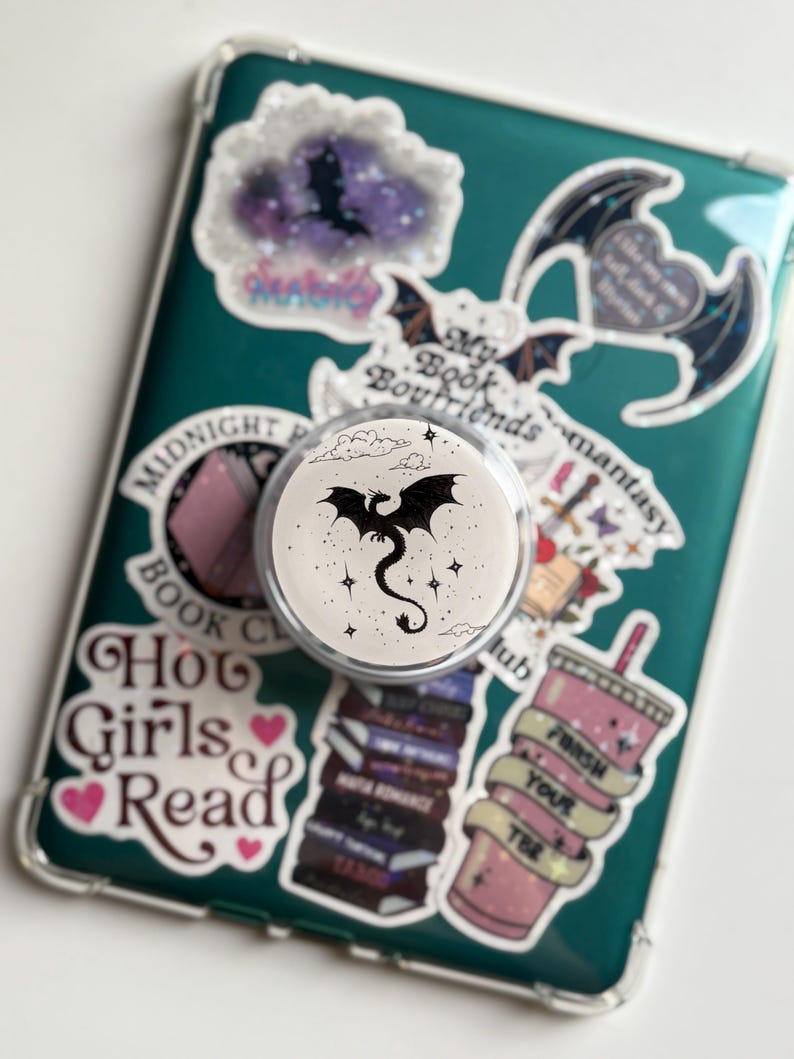 May include: A white PopSocket with a black dragon design, surrounded by stars, is attached to a teal phone case covered in stickers. The stickers feature bats, books, and text like "Hot Girls Read" and "My Book Boyfriends."