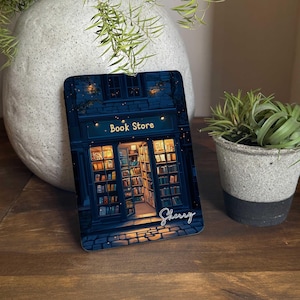 Könnte beinhalten: Eine nächtliche Illustration eines Buchladens mit den Worten "Book Store" über dem Eingang. Der Laden ist mit warmen gelben Lichtern beleuchtet und hat Regale voller Bücher. Das Bild hat ein blaues und schwarzes Farbschema mit einer weißen Signatur unten, die "Sherry" lautet.